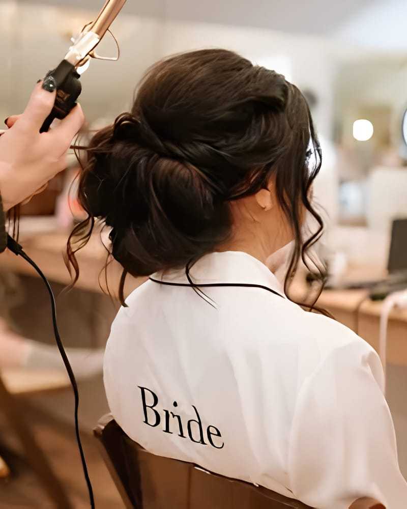 Hair stylist curling bride's hair in salon, wearing a robe with "Bride" on the back.