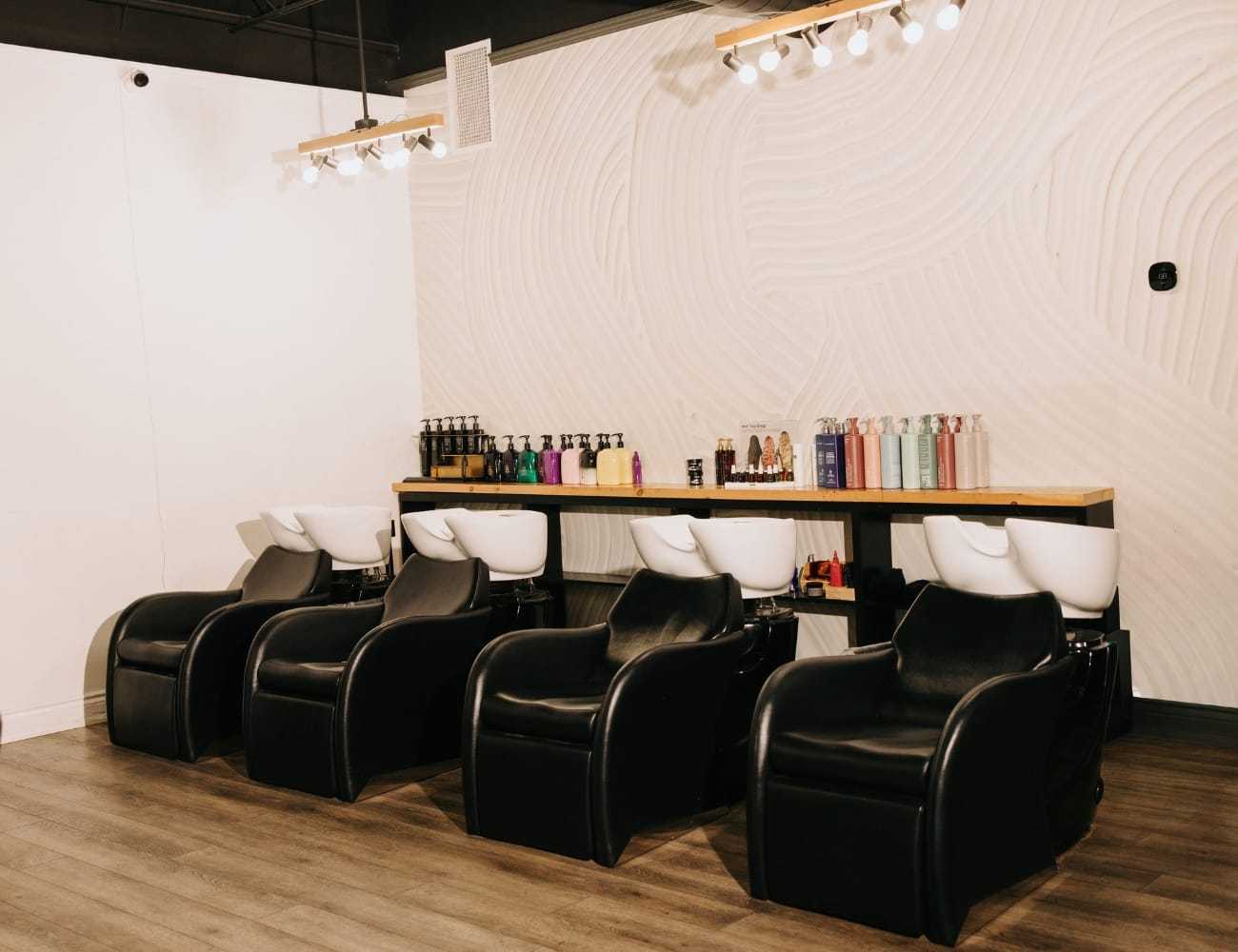 Four empty salon chairs positioned by hair-washing sinks with products on a wooden shelf.