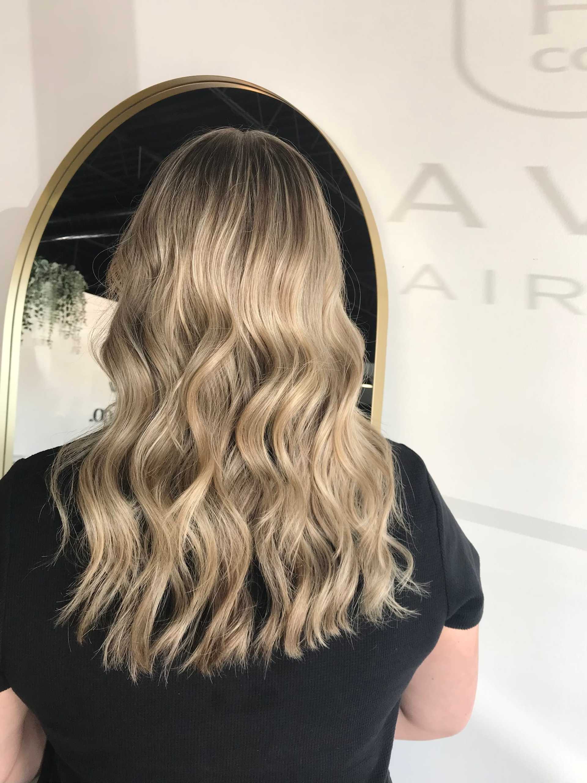 Wavy blonde hair styled in soft curls, viewed from behind, with a round mirror in the background.