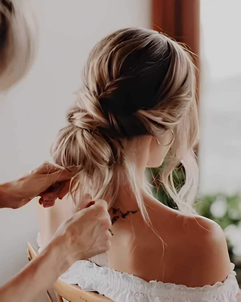 Hair being styled into an elegant bun, woman wearing an off-shoulder top, delicate tattoo visible.