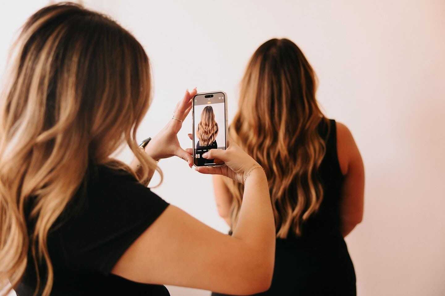 Two women in a salon: one taking a photo of the other's styled wavy hair with a phone.