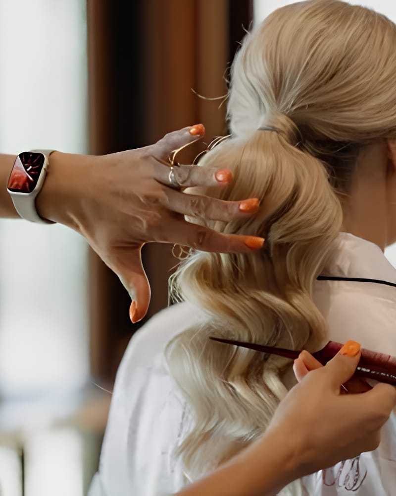 Woman getting her blonde hair styled with a curling iron; stylist is wearing a smartwatch.
